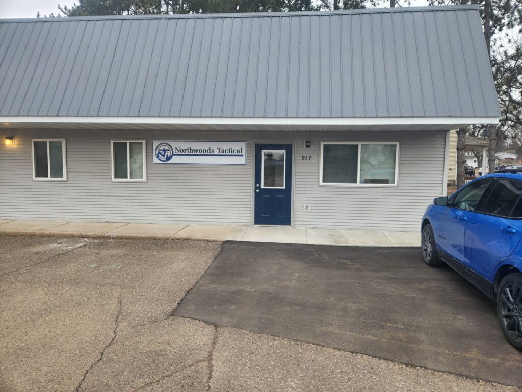 Front view of a gray building with a "Northwoods Tactical" sign, blue door, and partial blue car visible on the right.